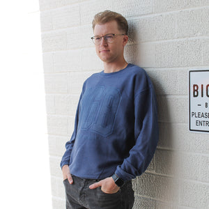 Man leaning against wall wearing a Big Grove Brewery University Puff Corded Crew 